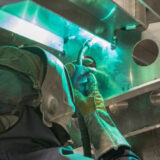 Worker in protective gear welding metal components inside a ship structure.
