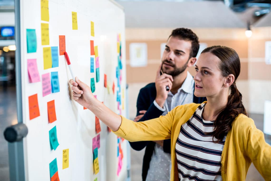 Woman reaching out to move a sticky note that is on a whiteboard with many other sticky notes, while a man looks on with a puzzled expression.