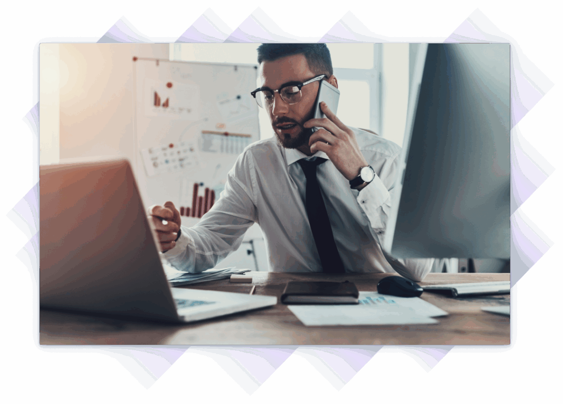 Very busy professional on a phone call while working on a laptop at a desk with charts and monitors.
