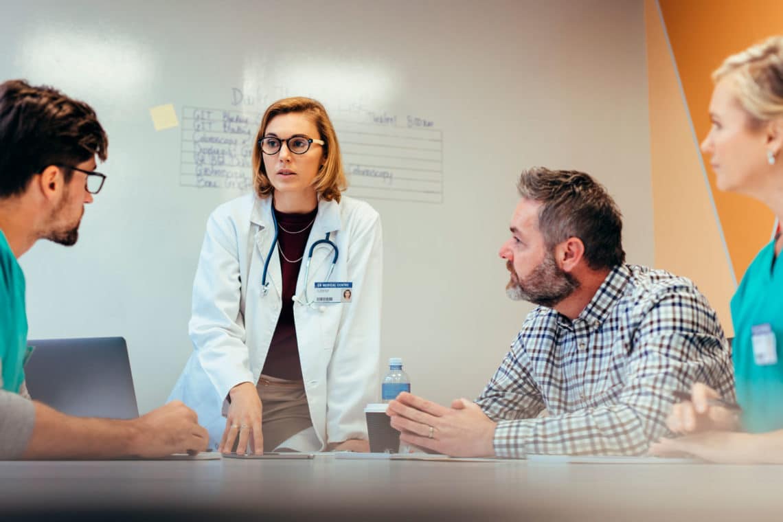Medical professionals in a room in front of a whiteboard having tense discussion.