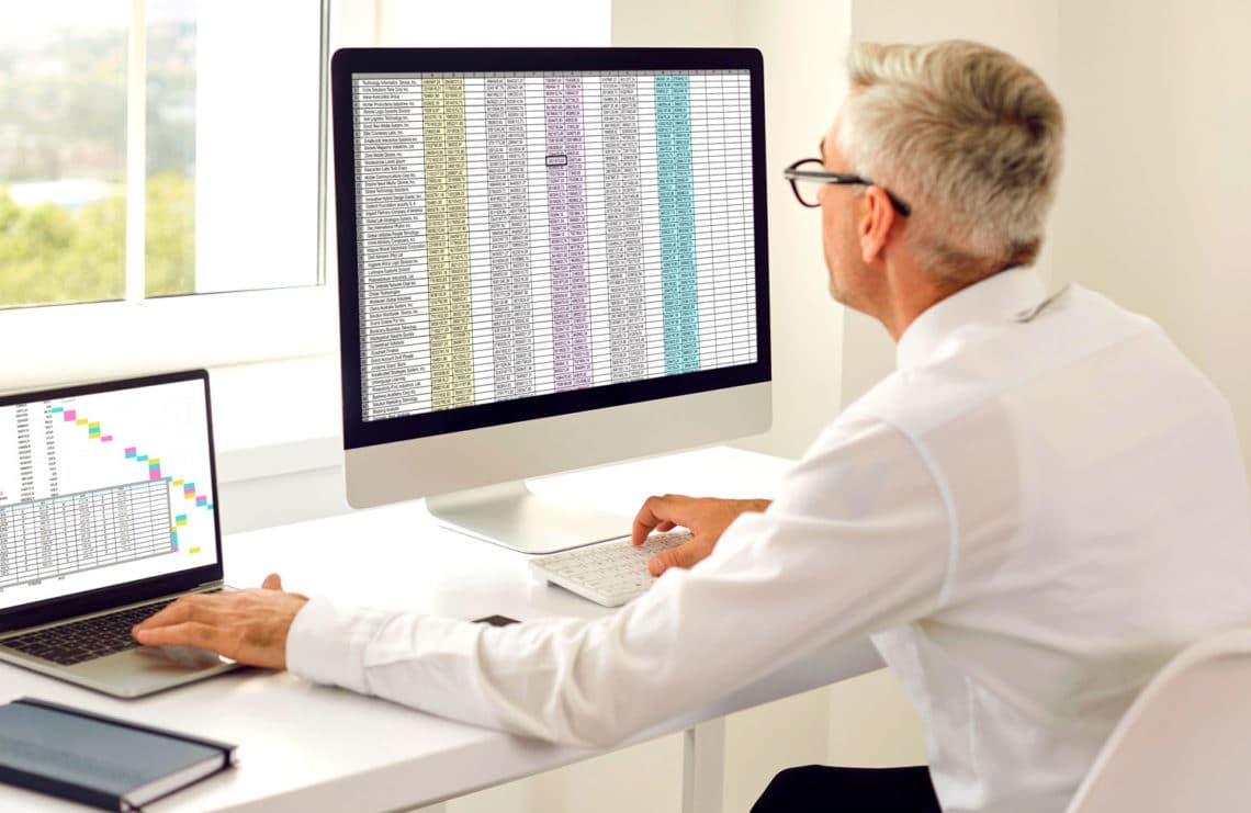 Man at desk in front of window looking at a large spreadsheet on monitor while comparing to another spreadsheet on a laptop.