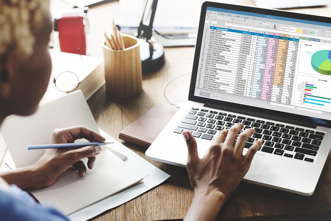 Woman writing notes on a notepad while reviewing pivot tables in Microsoft Excel on her laptop.