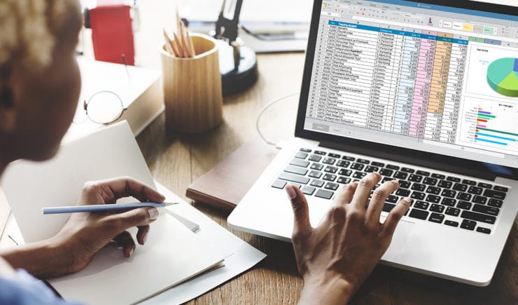 Woman writing notes on a notepad while reviewing pivot tables in Microsoft Excel on her laptop.