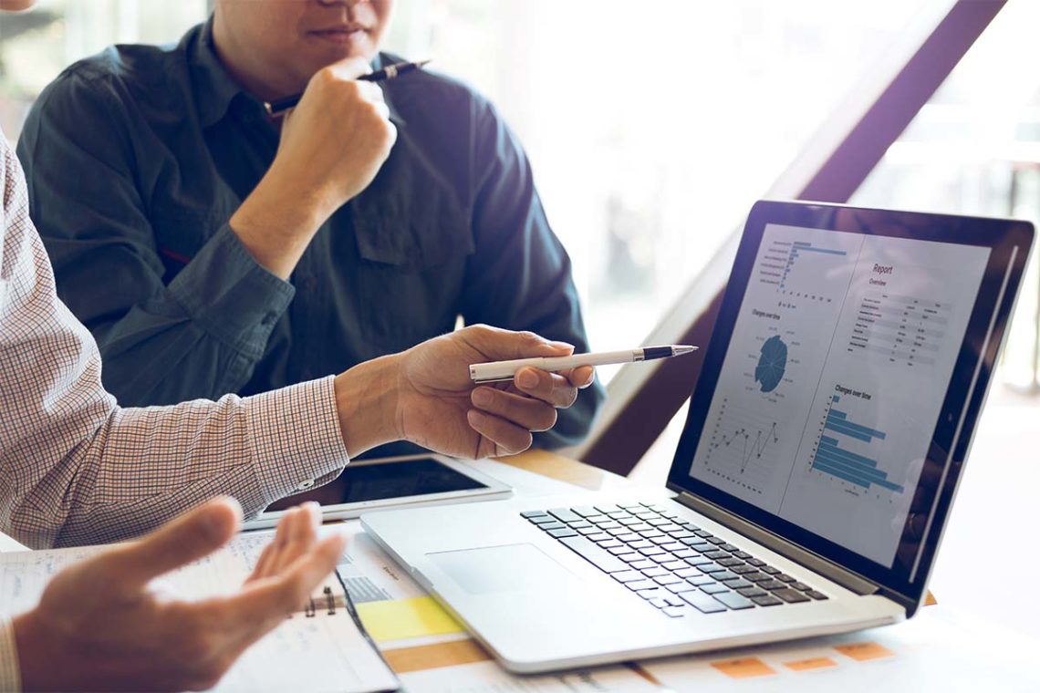 Man gesturing at a report with charts on laptop while his colleague considers the data.