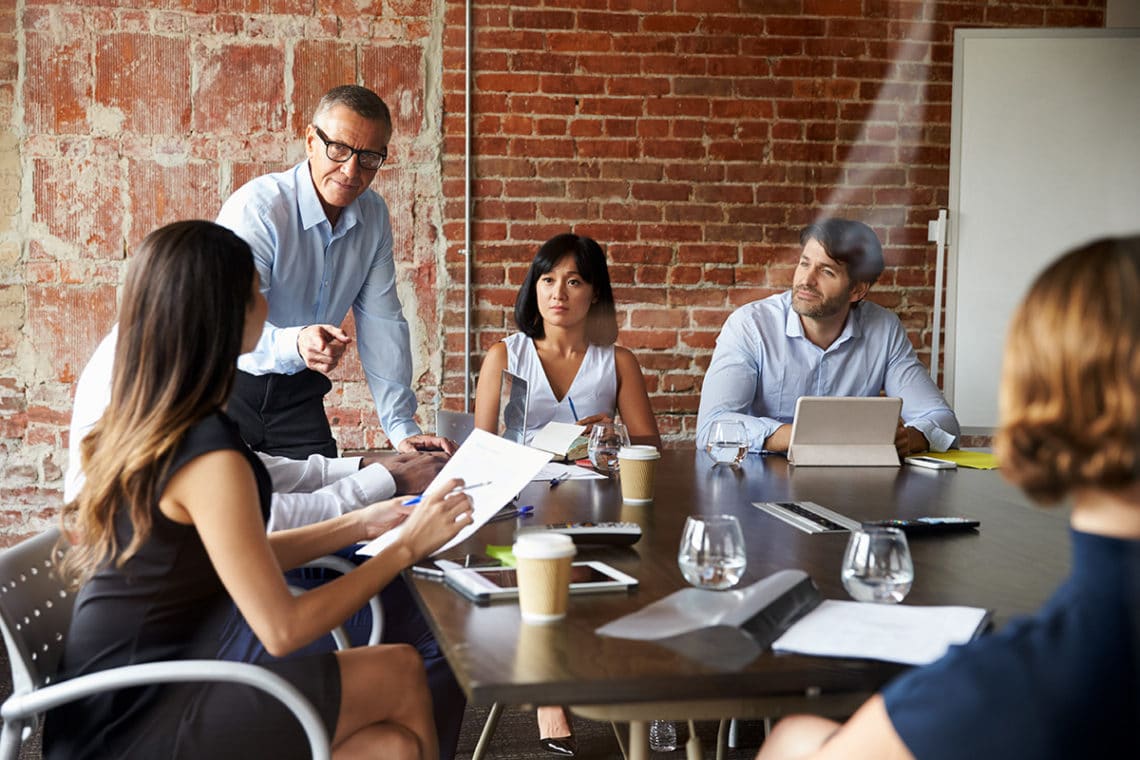 Man standing in a meeting room with his colleagues gesturing at a women holding reports.