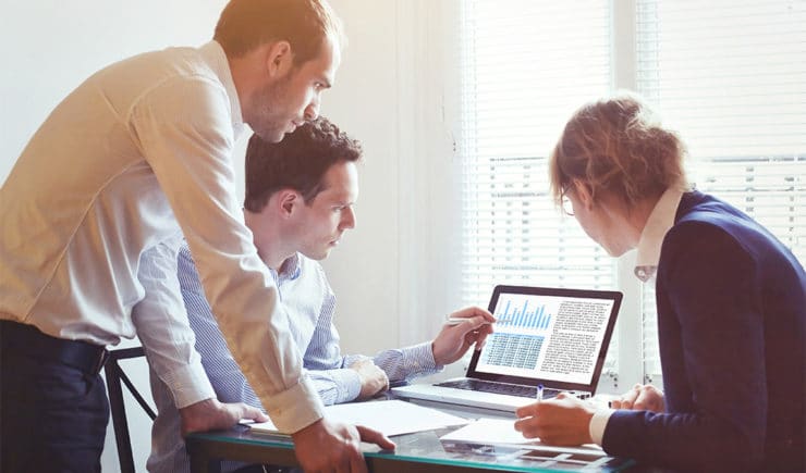 Several colleagues reviewing data on a laptop on a desk, while one points out an interesting aspect of the data.