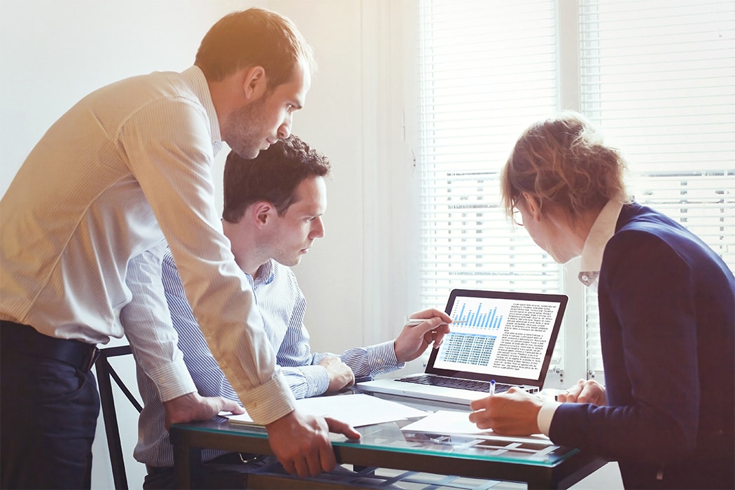 Several colleagues reviewing data on a laptop on a desk, while one points out an interesting aspect of the data.