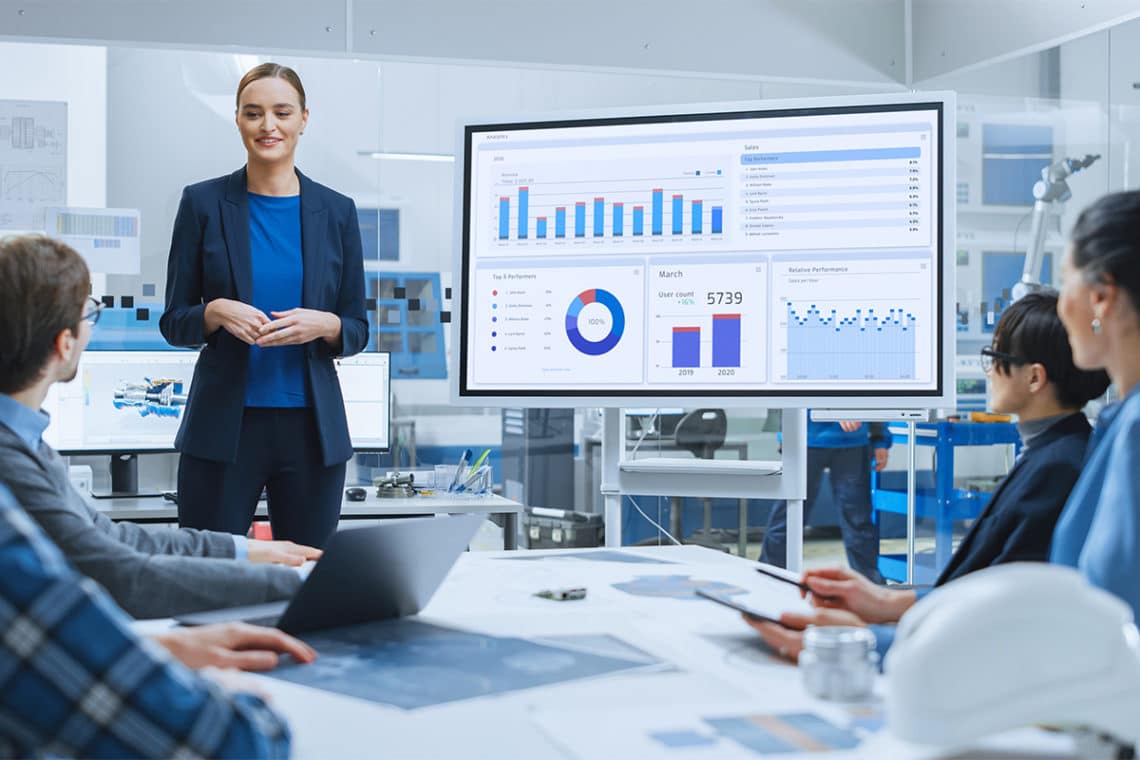 Woman presenting a full report on a large monitor to a group of colleagues who are watching intently.