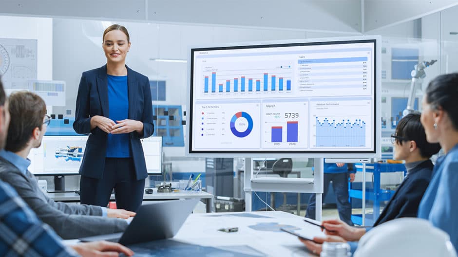 Woman presenting a full report on a large monitor to a group of colleagues who are watching intently.