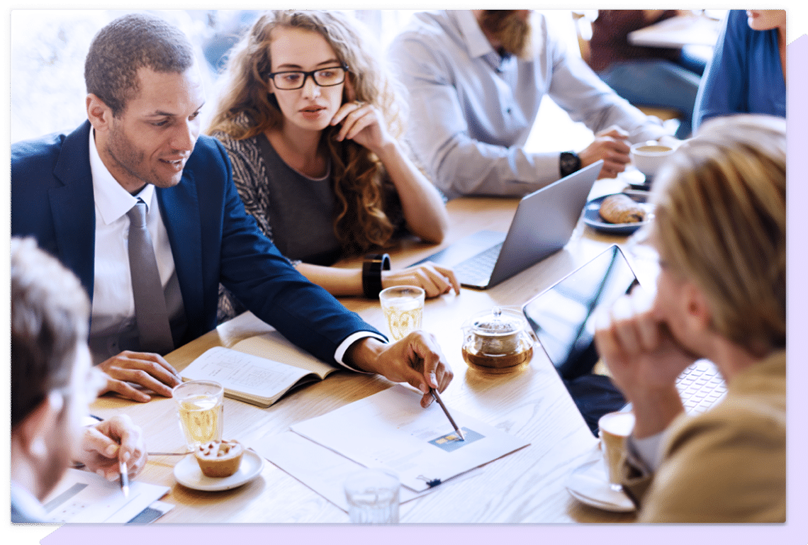 Team in a meeting around a large table where one man is pointing at a piece of paper with a pen.