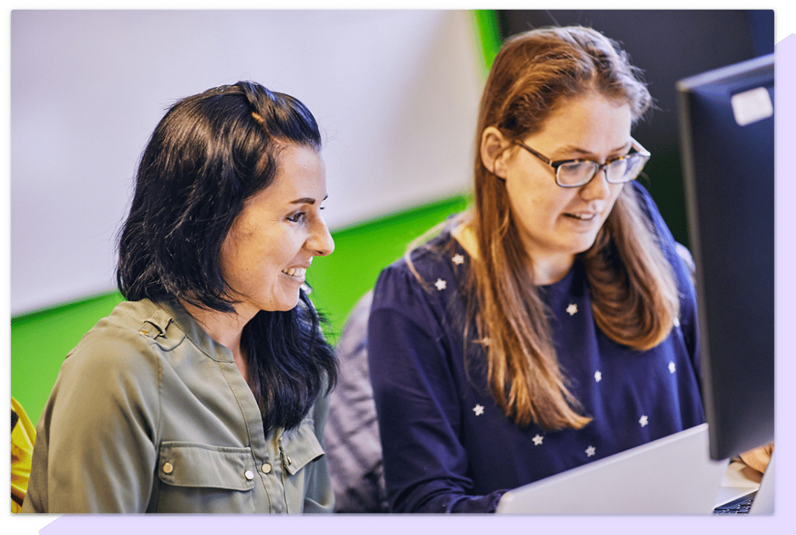 Two women looking at paperwork in front of a computer monitor.