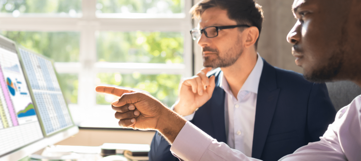 Two business professionals looking at spreadsheets and charts on two monitors.