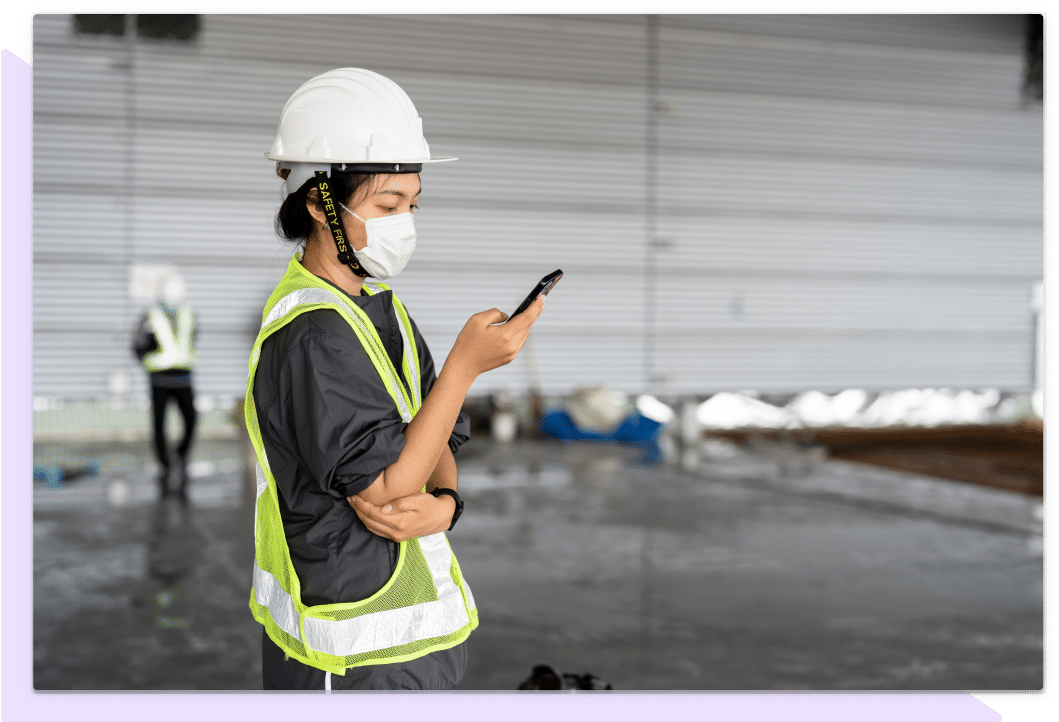 Employee in a white hard hat in face mask reviews information on a mobile phone.