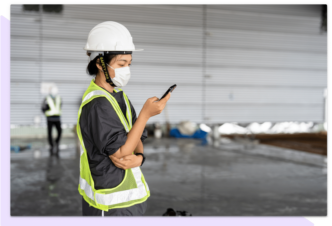 Employee in a white hard hat in face mask reviews information on a mobile phone.