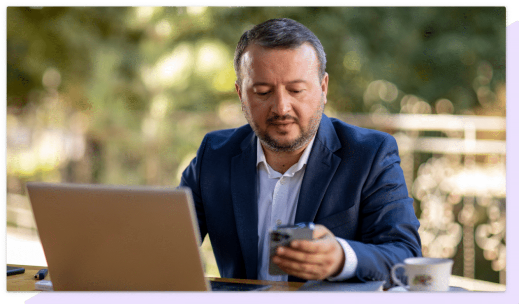 A businessman checks his phone while using a laptop in an outdoor setting.