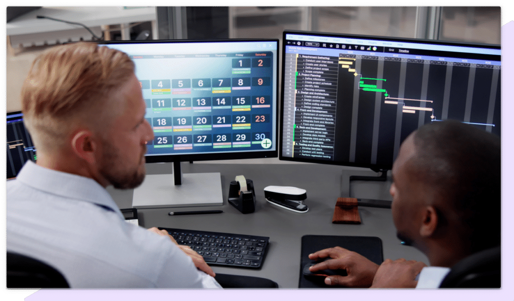 Two businessmen examine and discuss data displayed on two computer monitors.