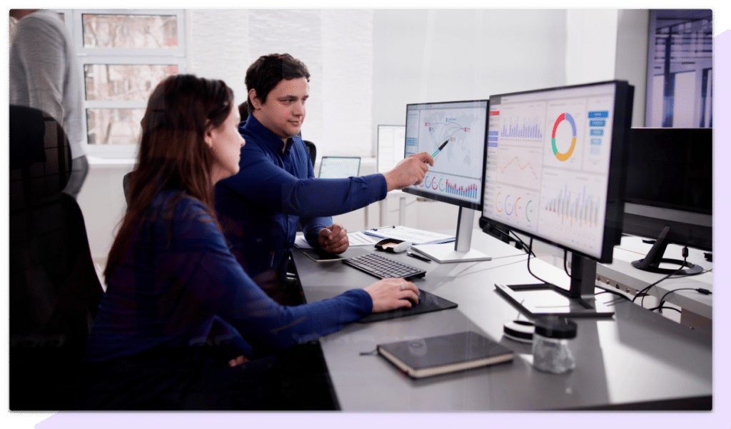 A businesswoman and man discuss a map and charts displayed on two computer monitors in an office.