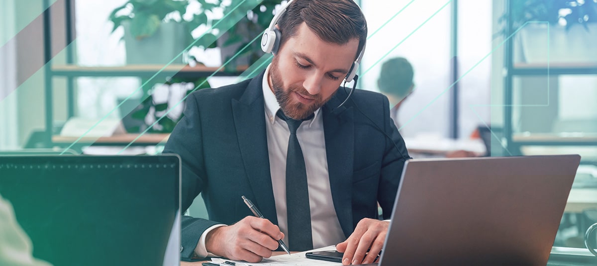 Businessman wearing headphones and taking notes with pen and paper in front of laptop.