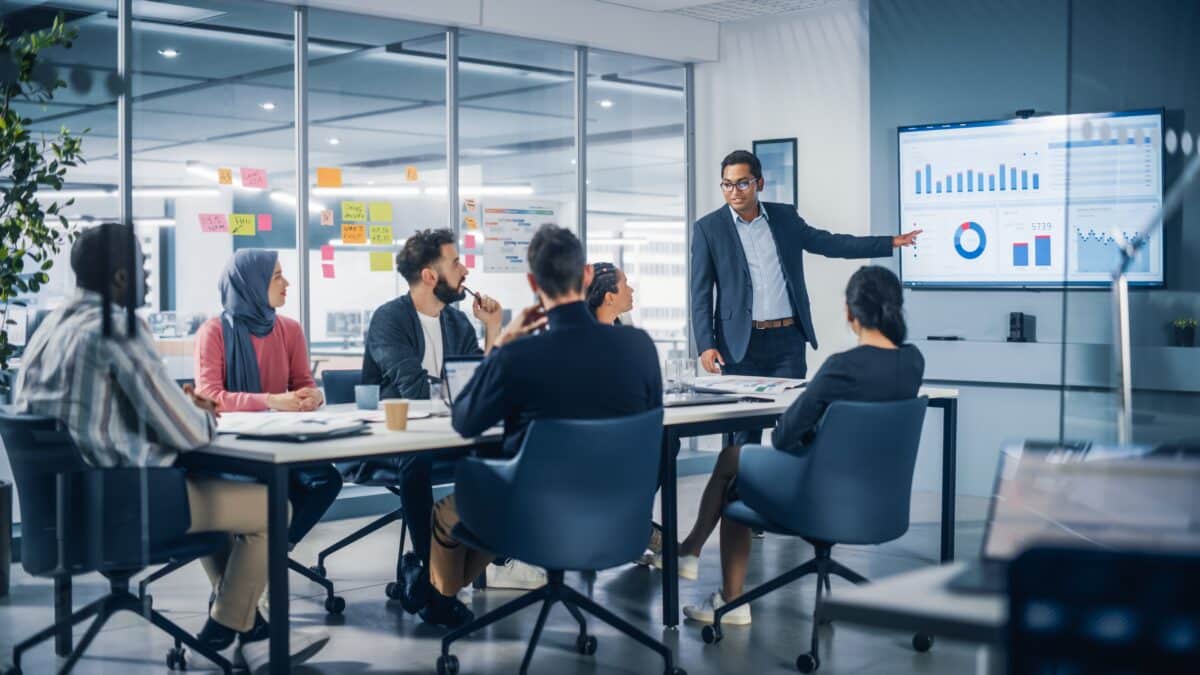 A trainer instructors a team of business professionals in an office, pointing to a whiteboard.