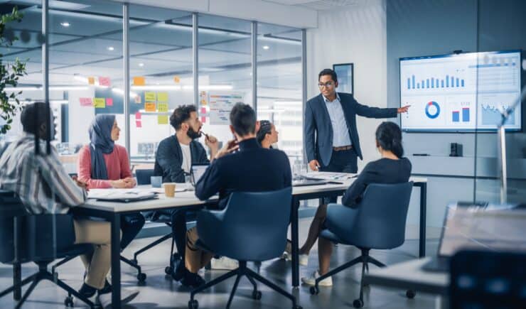 A trainer instructors a team of business professionals in an office, pointing to a whiteboard.