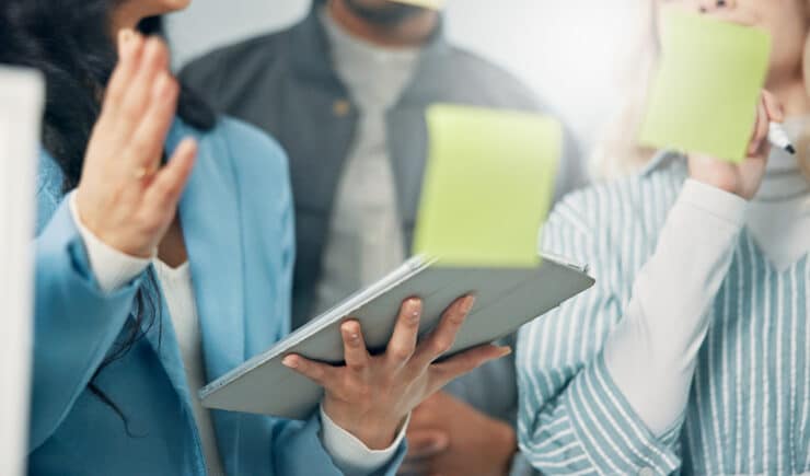 A group of people having a discussion, one person holding a tablet and another writing on a sticky note. The focus is on their hands and device.