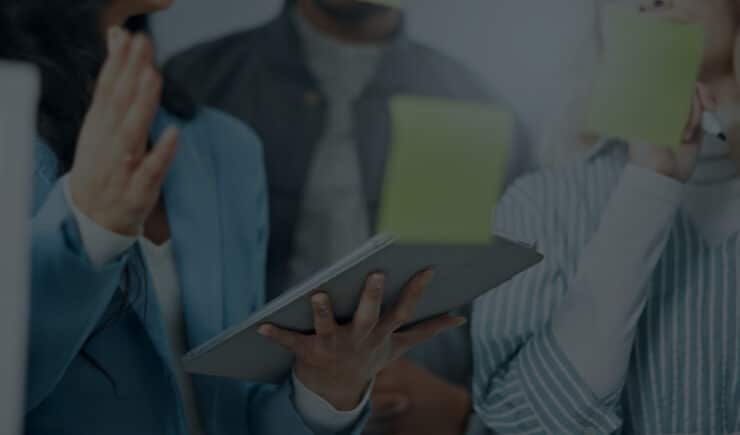 A group of people having a discussion, one person holding a tablet and another writing on a sticky note. The focus is on their hands and device.