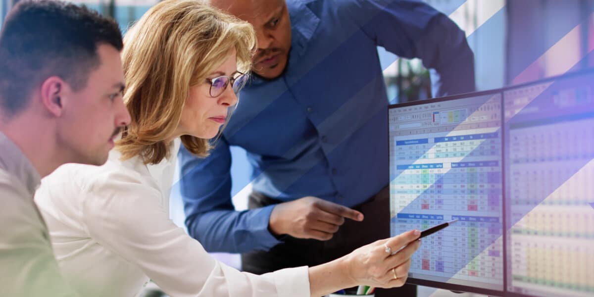 Three professionals focus intently on a computer monitor displaying complex spreadsheets, as one person points at the screen with a pen.
