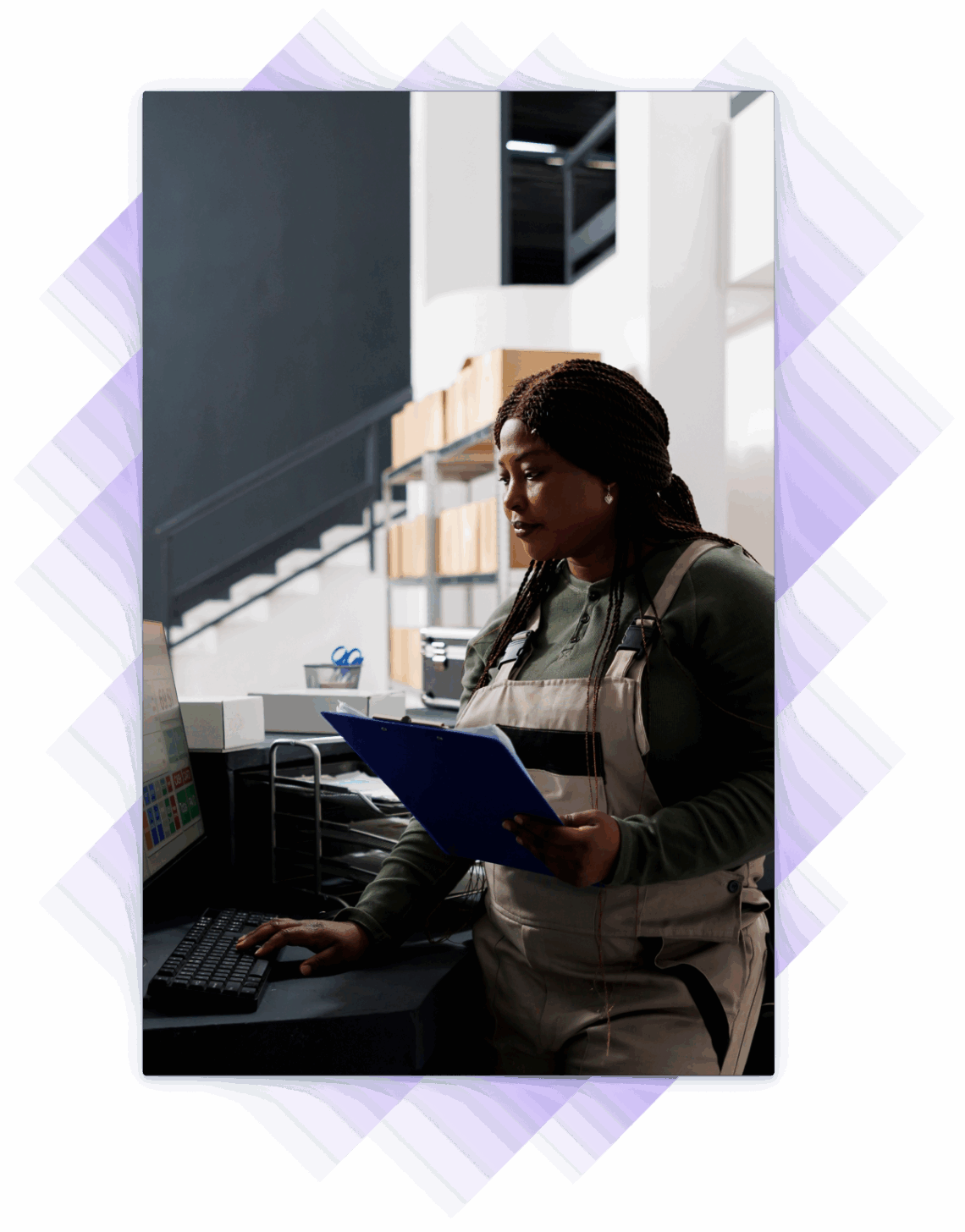 Woman in work overalls using a desktop computer while holding a clipboard in a stockroom.