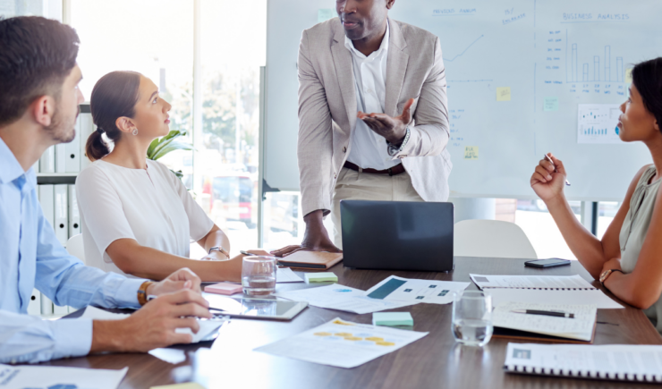 A business professional leads a meeting with four colleagues in a modern office, discussing data and strategy in front of a whiteboard with charts