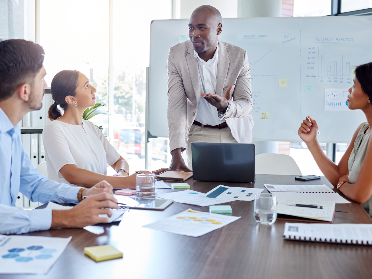 A business professional leads a meeting with four colleagues in a modern office, discussing data and strategy in front of a whiteboard with charts