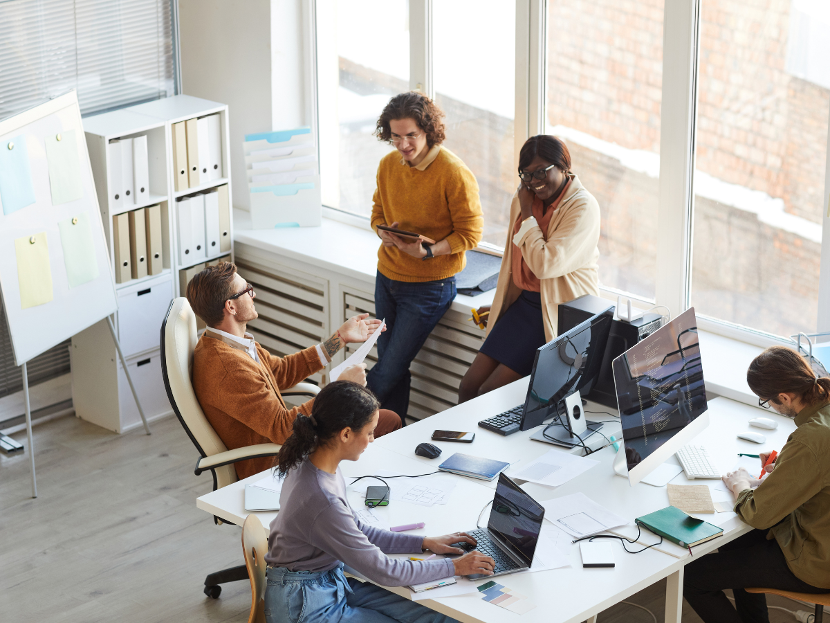 A diverse group of professionals collaborating in a modern office, reviewing charts and documents related to training management system features; two team members discuss near the window while others work at a shared desk.