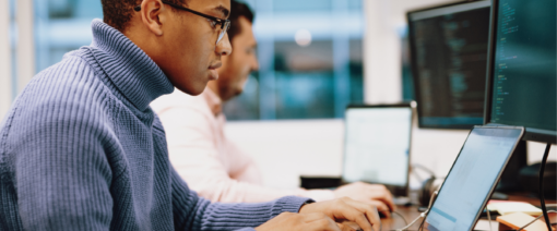 Focused professionals working at laptops and desktop monitors in a modern office, analyzing code or technical data.