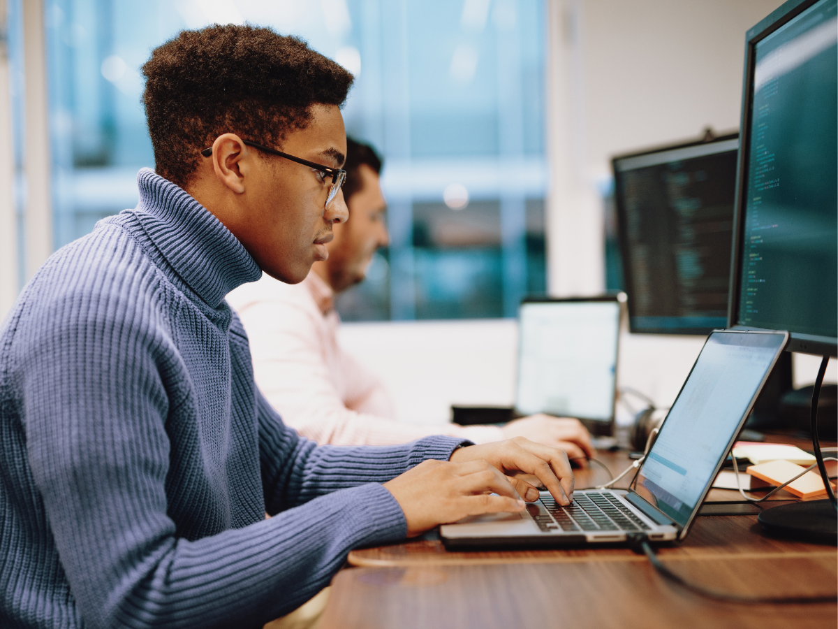 Focused professionals working at laptops and desktop monitors in a modern office, analyzing code or technical data.