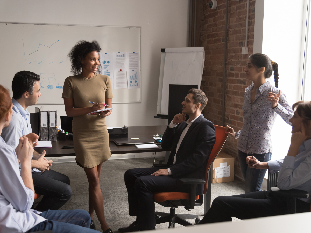 Smiling woman leading a casual office meeting with diverse team members, discussing charts and data in a modern conference room.