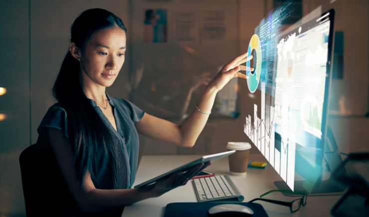 Woman analyzing data visualizations related to enterprise training success stories on a futuristic touchscreen, holding a tablet in a dark office.