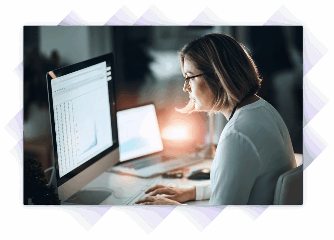 Woman looking closely at two monitors as the sun goes down.