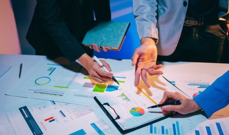Three people's hands point at and highlight various colorful bar charts, pie graphs, and data reports spread across a desk, illustrating a collaborative team review of performance or compliance metrics.
