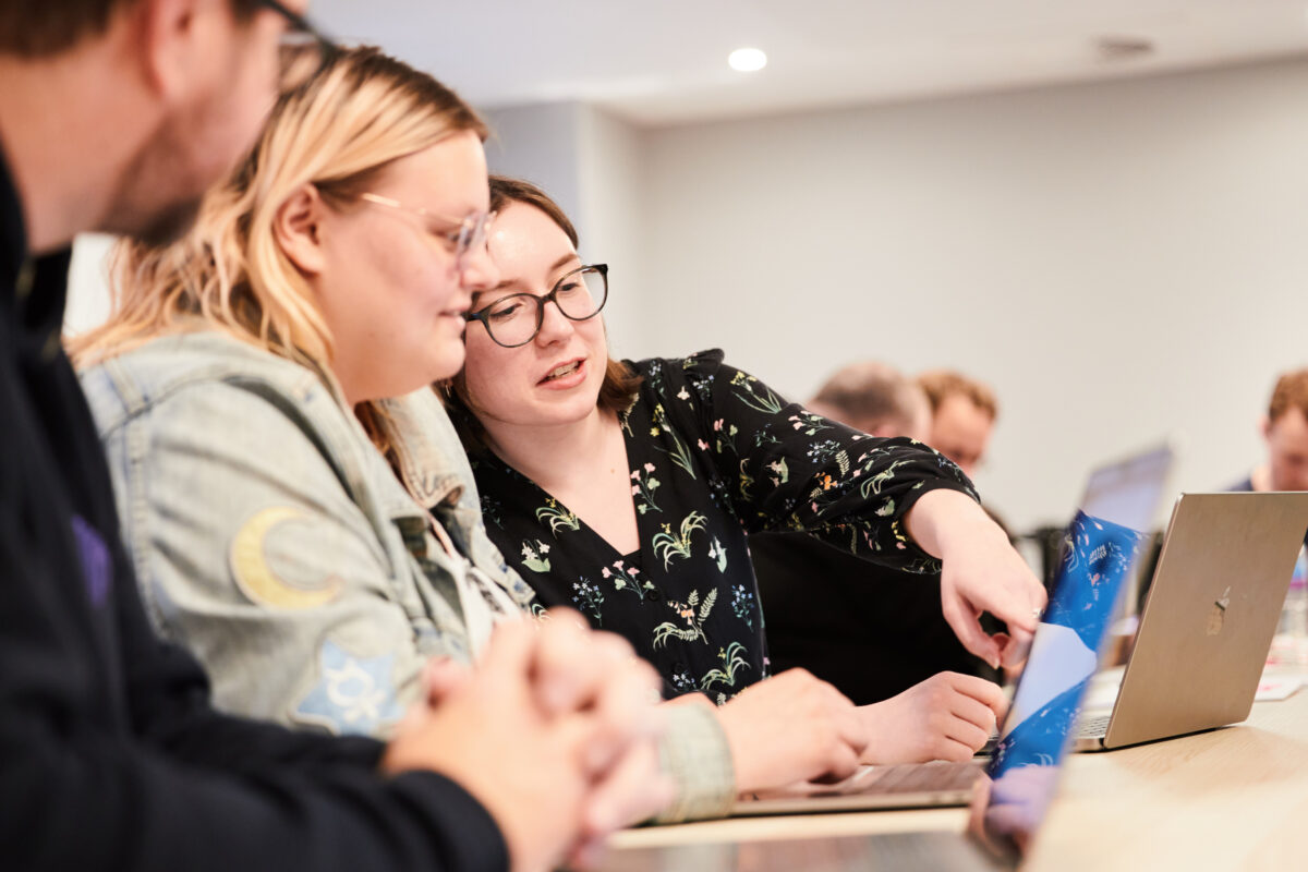 Two Administrate software engineers sitting around a laptop while one instructs the other by pointing at the screen.